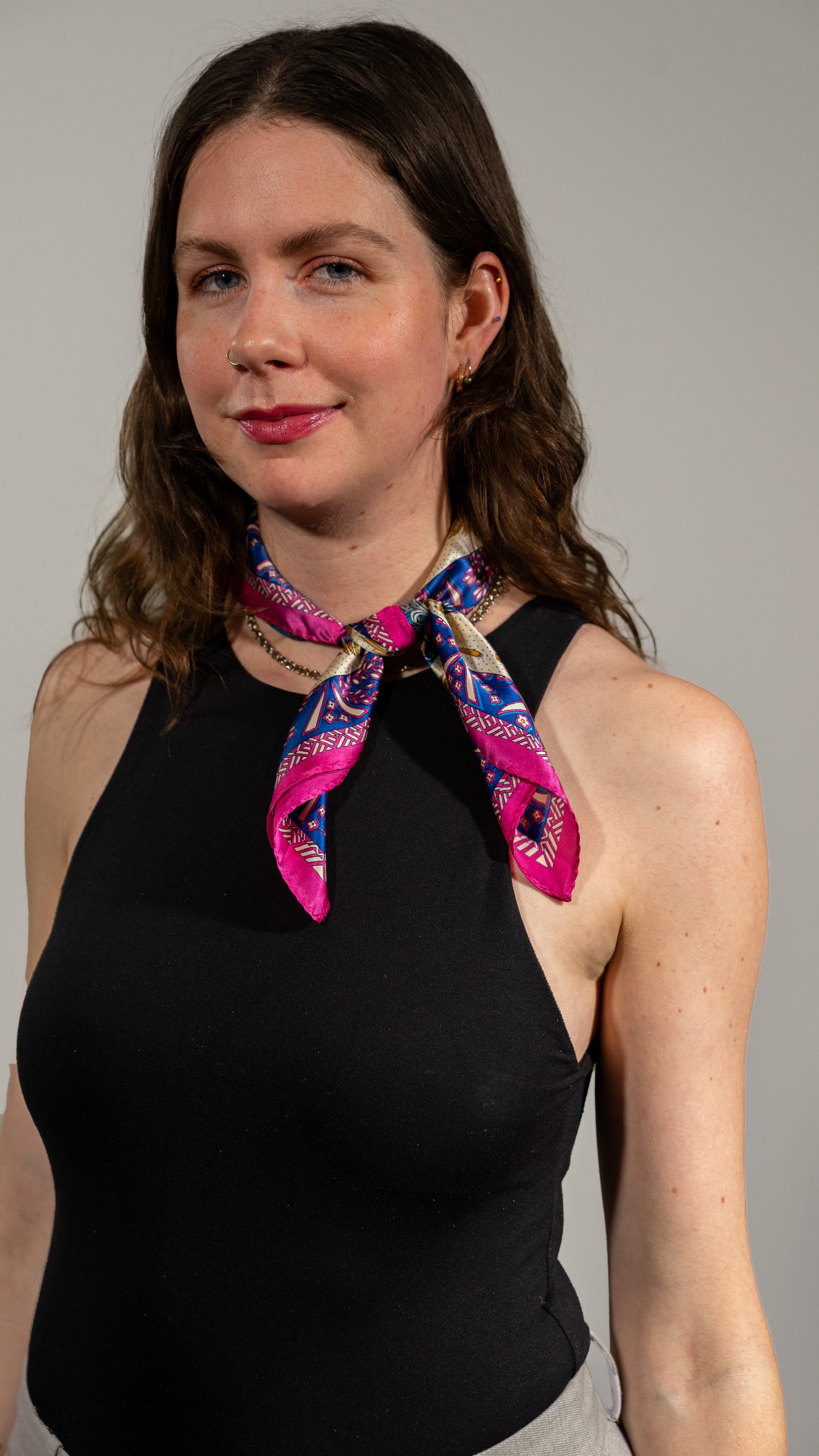 a female model wearing a square handwoven 100% pure silk scarf with vibrant blue, purple, and pink colours and intricate designs lightly tied around her neck, looking at the camera.