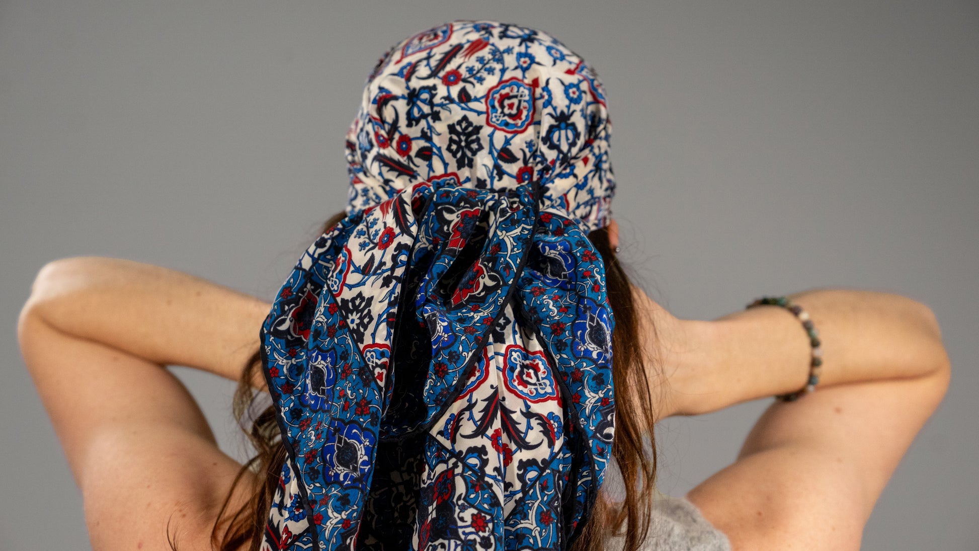 model wearing a vegan silk scarf with blue, red, black and white tones, featuring intricate patterns, styled as a hair wrap. Back-side angle