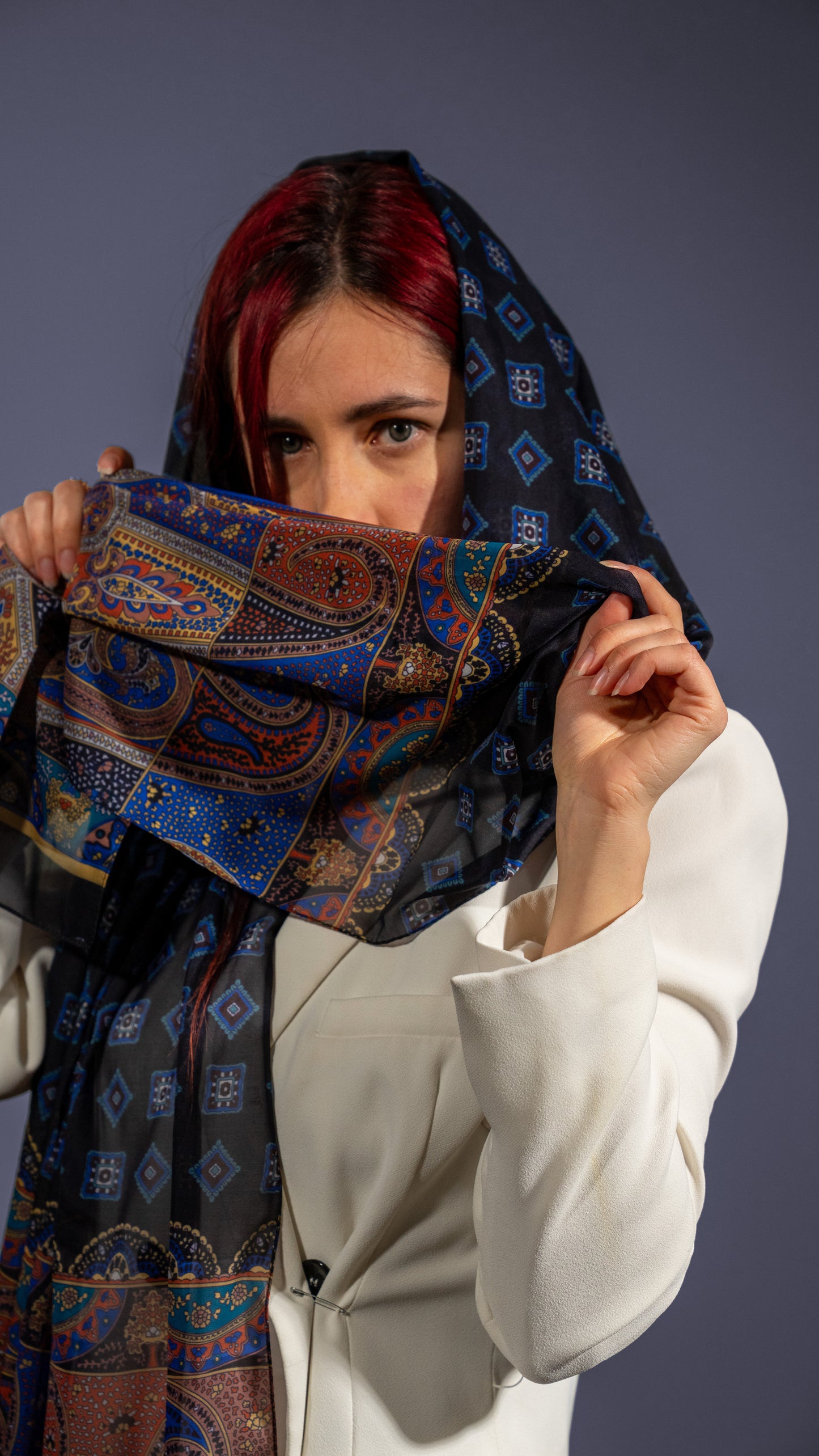 female model in a white suit wearing a colourful cotton voile shawl with intricate designs, featuring black, blue, and orange tones. Styled as a hair wrap, holding it up with hands