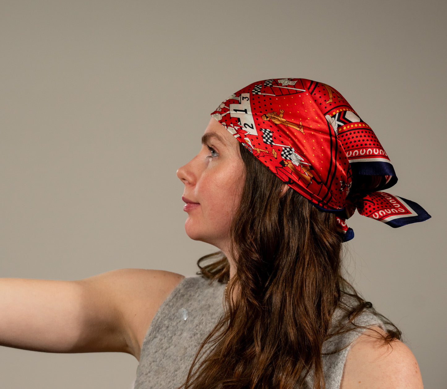 female model wearing square silk scarf, 100% pure silk, design with humans and race horses, predominantly red and white tones, styled as a head wrap. Side angle.