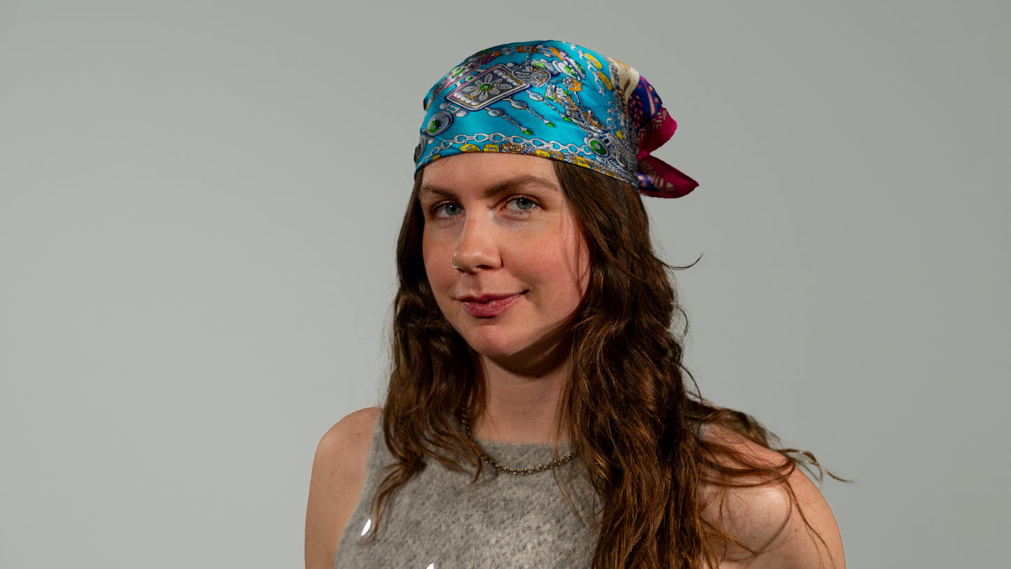 a female model wearing a square handwoven 100% pure silk scarf with vibrant blue, purple, and pink colours and intricate designs as a hair wrap, smiling at the camera.