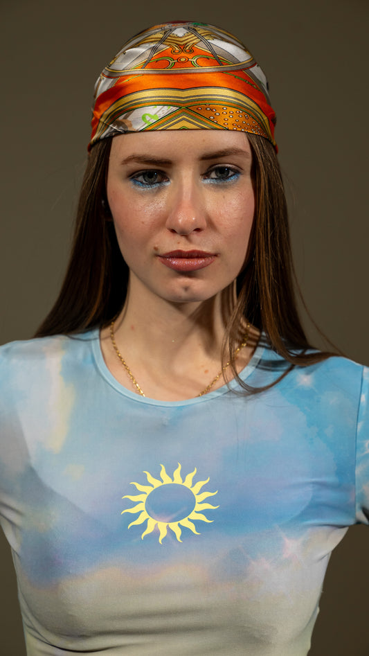 a female model wearing a colourful square 100% pure silk scarf, featuring circular designs with a predominantly orange colour as a headwrap.